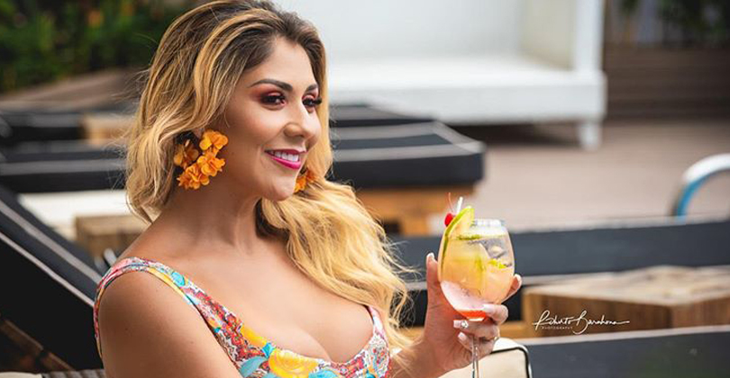 Picture of a female guest sitting next to the pool of the Costa Rica Medical Center Inn, San Jose, Costa Rica.  The guest has a flower in her sandy blonde hair and is smiling as she is holding a tropical drink with her hand.