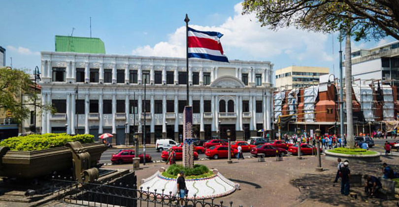 Picture of a park and museum in the city, illustrating the sightseeing attractions available for patients of Frontline Plastic Surgery in San Jose, Costa Rica.  The picture shows a large building with taxis parked in front and a large Costa Rican flag on a flag pole in the center of the park.
