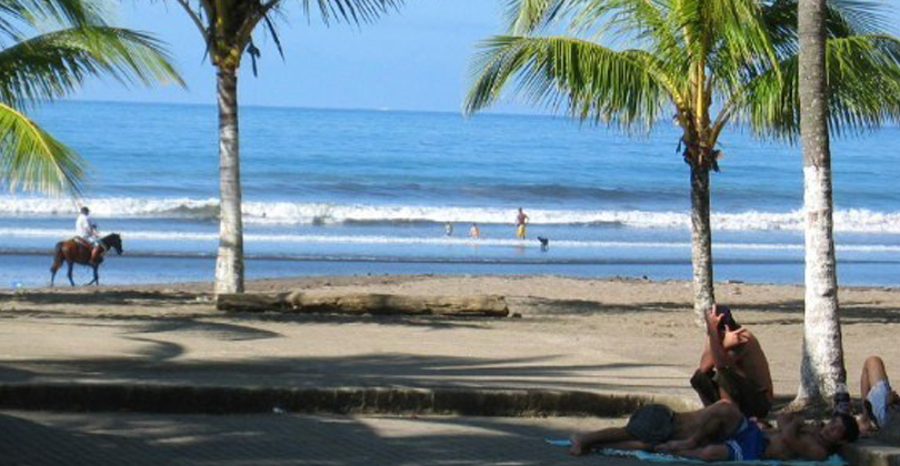 Picture of a beautiful sandy beach with palm trees, representing the sightseeing opportunities while having plastic surgery with Frontline Dental CR, San Jose, Costa Rica.  The water is deep blue with gently breaking waves and a horseback rider is shown on the seashore.