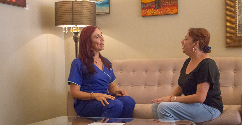 Picture of Estela Araya, International Coordinator of the Costa Rica Medical Center Inn, San Jose, Costa Rica sitting with a guest.  Estela is wearing a blue uniform and is sitting with the guest on a beige couch in the lobby.