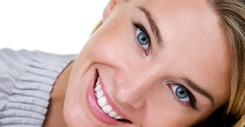 Picture of a beautiful female patient, happy with the dental crowns procedure she had at Frontline Dental CR in San Jose, Costa Rica.  The picture shows a woman with brown hair, holding her hand under her cheek, and smiling while looking directly at the camera.