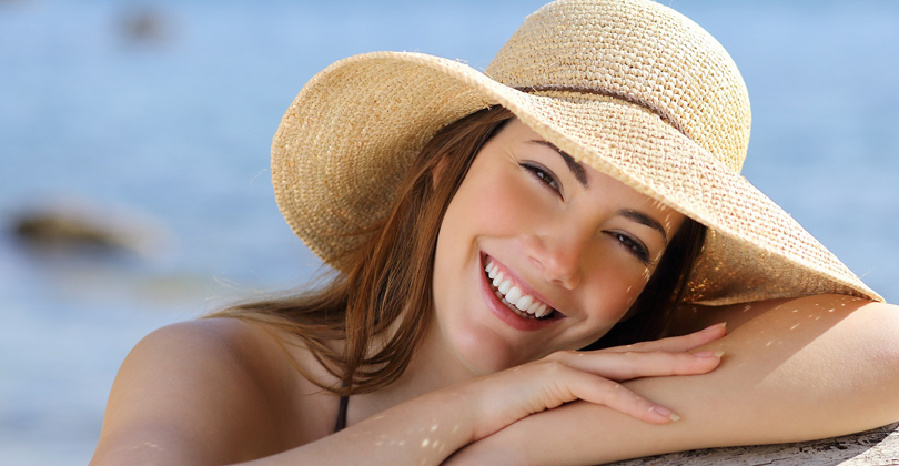 Picture of a female patient, happy with the dental implants she had at Frontline Dental CR in San Jose, Costa Rica. The picture shows a beautiful smiling woman wearing a straw hat with her arms folded and smiling directly at the camera.
