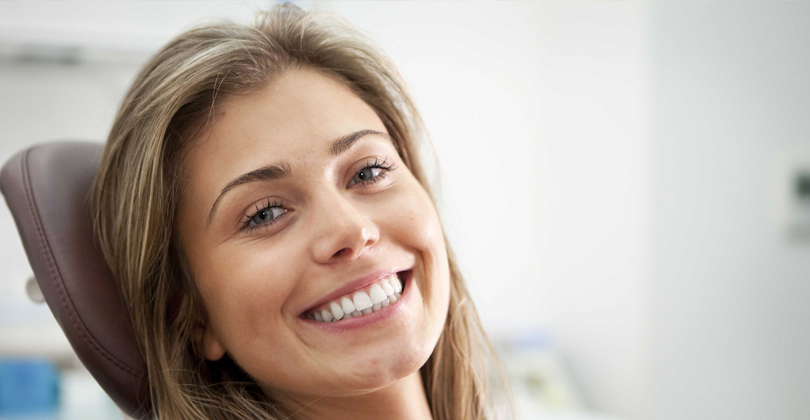 Picture of a beautiful female patient, happy with the dentures she had at Frontline Dental CR in San Jose, Costa Rica.  The picture shows a woman with long sandy brown hair, sitting in a dental chair, and smiling while looking directly at the camera.