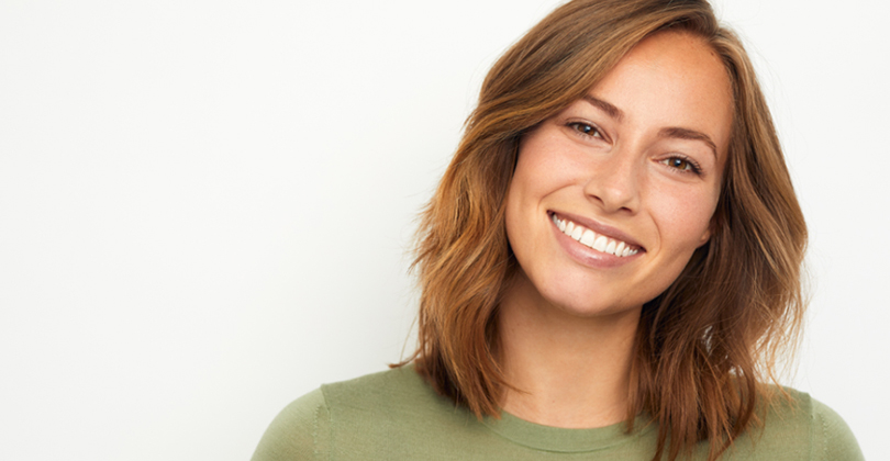 Picture of a beautiful female patient, happy with the full mouth dental makeover she had at Frontline Dental CR in San Jose, Costa Rica.  The picture shows a woman with long brown hair, tilting her head to the side and smiling while looking directly at the camera.