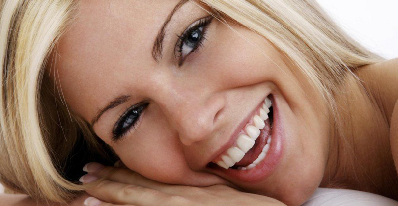 Picture of a beautiful female patient, happy with the Hollywood caps she had at Frontline Dental CR in San Jose, Costa Rica.  The picture shows a woman with blonde hair, holding her hand to the side of her face, and smiling while looking directly at the camera.