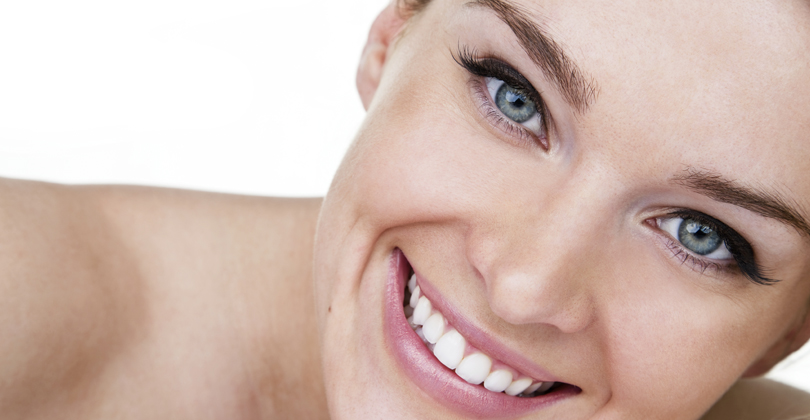 Picture of a beautiful female patient, happy with the implant-supported bridge she had at Frontline Dental CR in San Jose, Costa Rica.  The picture shows a woman with short brown hair, with one hand to the side of her face, and smiling while looking directly at the camera.