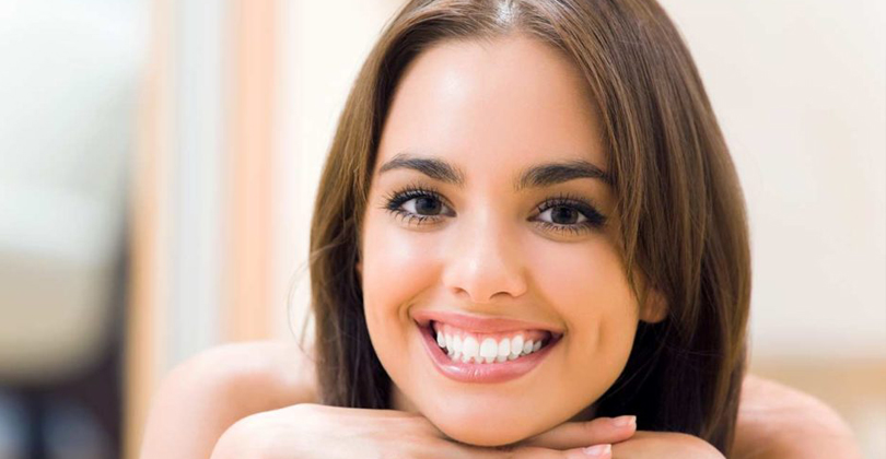 Picture of a beautiful female patient, happy with the implant-supported dentures she had at Frontline Dental CR in San Jose, Costa Rica.  The picture shows a woman with long brown hair, holding both hands under her chin, and smiling while looking directly at the camera.