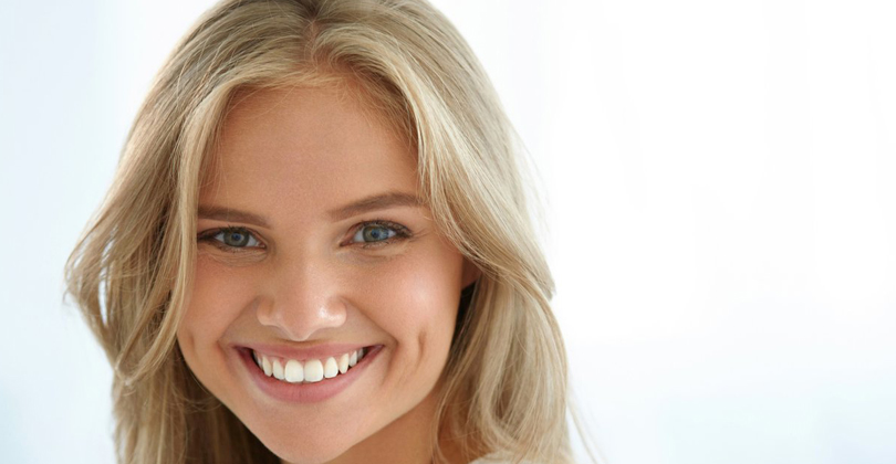 Picture of a beautiful female patient, happy with the periodontal treatment she had at Frontline Dental CR in San Jose, Costa Rica.  The picture shows a woman with long blonde hair, wearing a white blouse, and smiling while looking directly at the camera.